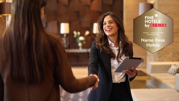 Eine Hotelmitarbeiterin begrüßt freundlich eine Gästin an der Rezeption, daneben das Fine Hotels by KURIER Gütesiegel als Auszeichnung für besondere Servicequalität.