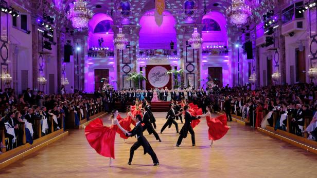 In einem prunkvollen Saal tanzen Paare in roter Ballkleidung und Frack vor einem Publikum.
