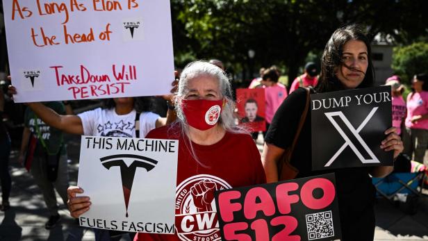 Mehrere Menschen halten Protestschilder mit kritischen Botschaften zu Tesla, X und Elon Musk bei einer Demonstration.