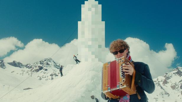 Mann mit Ziehharmonika vor verpixelter Schneeskulptur in winterlicher Berglandschaft.
