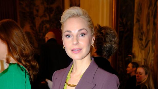 Eine Frau mit blondem Haar trägt einen lila Hosenanzug mit grünem Hemd und hält eine schwarze Handtasche.