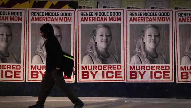 Eine Person mit Kapuze geht an mehreren Plakaten vorbei, die das Porträt einer Frau und den Text „Murdered by ICE“ zeigen.
