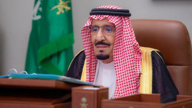 König Salman von Saudi-Arabien sitzt an einem Tisch mit der saudischen Flagge im Hintergrund.