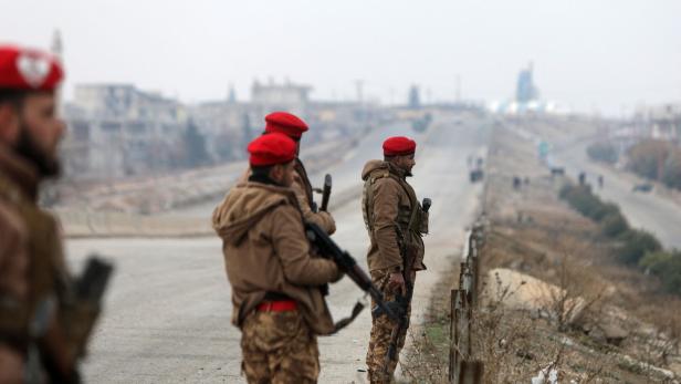 Soldaten mit roten Baretts und Gewehren stehen auf einer Straße vor einer zerstörten Stadt.