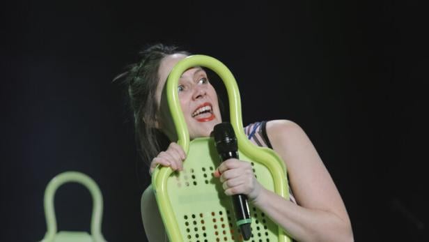 Frau in glitzerndem Kleid hält ein Mikrofon und einen grünen Stuhl, während sie auf einer Bühne steht.