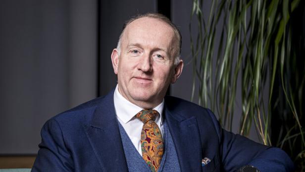 Ein Mann im Anzug mit bunter Krawatte sitzt auf einem grünen Sessel, im Hintergrund steht eine große Pflanze.