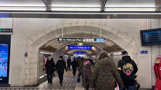Menschen gehen durch einen Tunnel mit gemustertem Boden und Hinweisschildern zu Bahnsteigen und Ausgängen.
