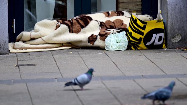 Eine Person liegt unter einer Decke auf dem Gehweg, daneben stehen eine Plastiktüte und eine Einkaufstasche, im Vordergrund Tauben.