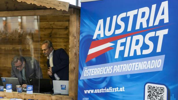 Zwei Männer arbeiten an Laptops in einem Holzstand neben einem "Austria First"-Banner.