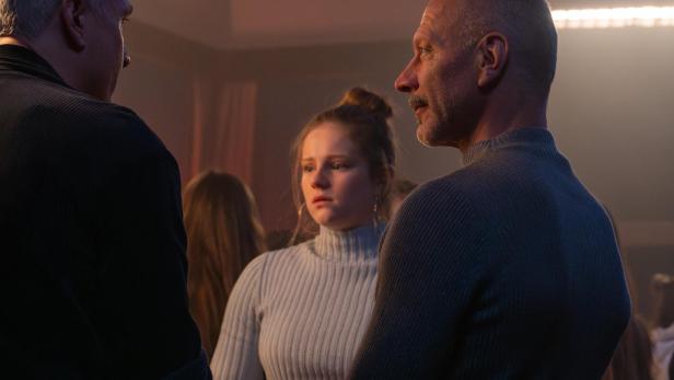 Drei Personen stehen in einem Raum, eine junge Frau in der Mitte mit besorgtem Blick.