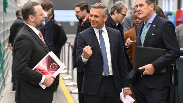 Drei Männer im Anzug unterhalten sich in einer modernen Halle, umgeben von weiteren Personen im Hintergrund.