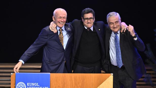 Drei Männer in Anzügen umarmen sich lachend vor einem Rednerpult der Königlich Schwedischen Akademie der Wissenschaften.