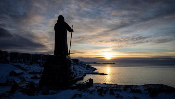 Eine Statue mit Stab steht auf einem Hügel im Schnee und blickt auf das Meer bei Sonnenuntergang.
