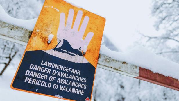 Warnschild mit erhobener Hand und Hinweis auf Lawinengefahr in mehreren Sprachen, umgeben von Schnee.