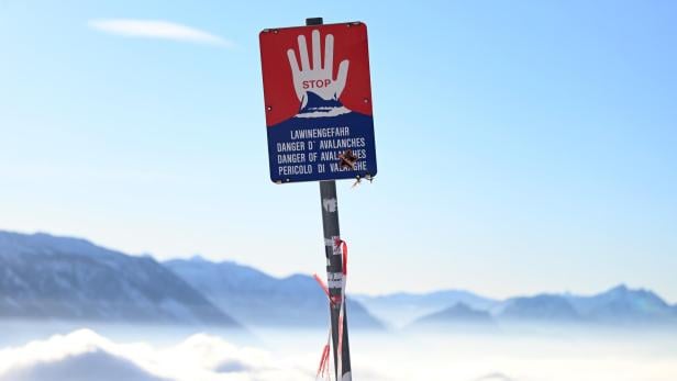Warnschild mit roter Hand und Hinweis auf Lawinengefahr vor verschneiten Bergen bei klarem Himmel.