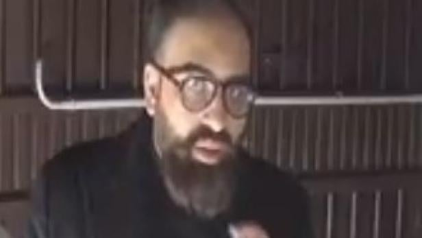 Mann mit Bart und Brille in dunkler Kleidung steht vor einer Holzwand.