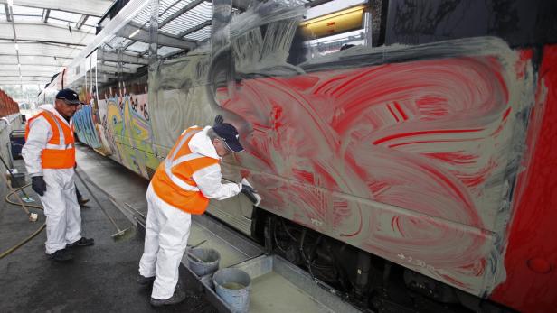 Zwei Personen in Schutzkleidung und Warnwesten reinigen Graffiti von einem Zug mit Bürsten und Reinigungsmitteln.