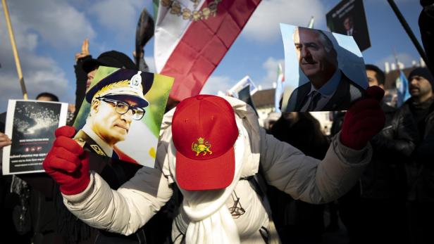 Eine Person mit roter Kappe hält Porträts zweier Männer, im Hintergrund eine Flagge und weitere Demonstrierende.