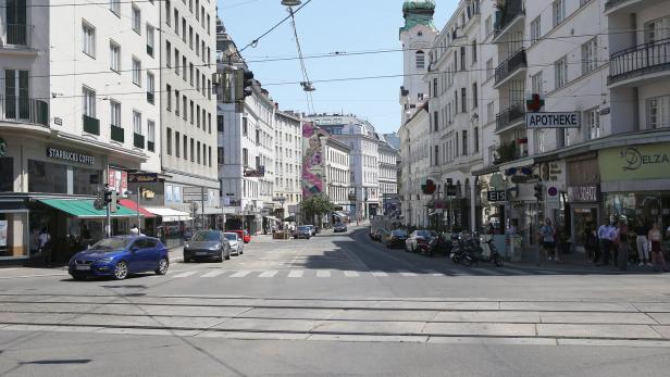 Mehrspurige Straße mit Autos, Straßenbahnschienen, Geschäften, Menschen auf Gehwegen und einer Kirche mit grünem Turm im Hintergrund.