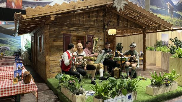 Vier Musiker in traditioneller Tracht spielen Blasinstrumente und Gitarre vor einer Holzhütte mit Alpenkulisse.