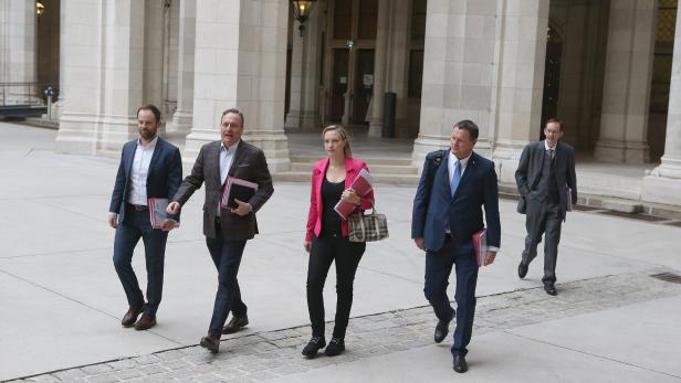 Fünf Personen, darunter Taborsky und Hungerländer,  in Businesskleidung gehen mit Aktenmappen durch einen historischen Arkadengang.