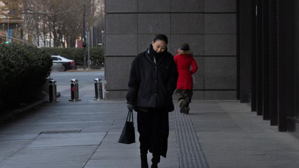 Eine Person in dunkler Kleidung und Maske geht auf einem Gehweg, im Hintergrund läuft jemand in rotem Mantel davon.