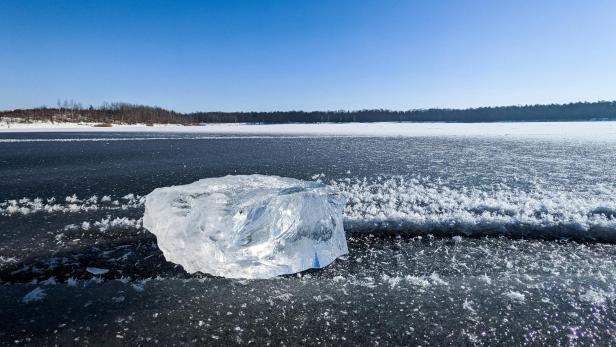 Große Eisscholle liegt auf einer zugefrorenen, schneebedeckten Fläche unter blauem Himmel.