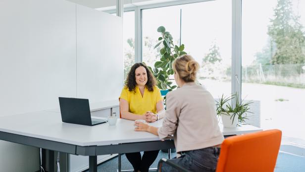 Zwei Frauen sitzen sich an einem Schreibtisch in einem hellen Büro gegenüber und unterhalten sich.