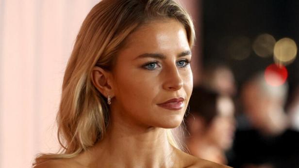 Caro Daur mit langen blonden Haaren trägt ein schulterfreies schwarzes Kleid und silbernen Schmuck, im Hintergrund unscharfe Personen.