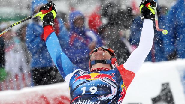 Ein Skirennläufer mit Startnummer 19 jubelt mit erhobenen Armen im Schneefall, im Hintergrund Zuschauer.