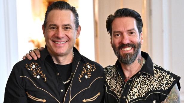 Zwei Männer mit bestickten schwarzen Hemden stehen lächelnd nebeneinander, einer hat sichtbare Tattoos am Arm.