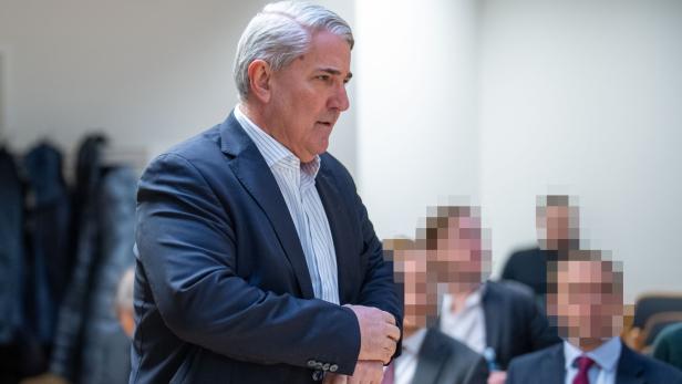 Ernst Nevrivy mit grauem Haar und Anzug steht vor einer Gruppe von Personen.