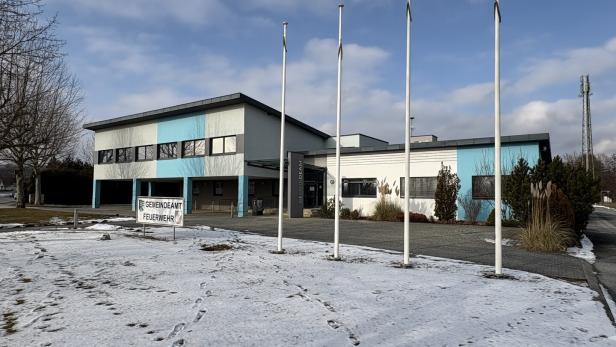 Ein Gebäude des Gemeindeamts/Feuerwehr mit vier Fahnenmasten steht vor einem teilweise schneebedeckten Feld.