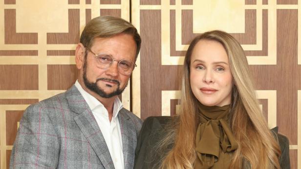 Ein Mann im karierten Anzug und eine Frau mit langen blonden Haaren und olivgrünem Outfit stehen nebeneinander.