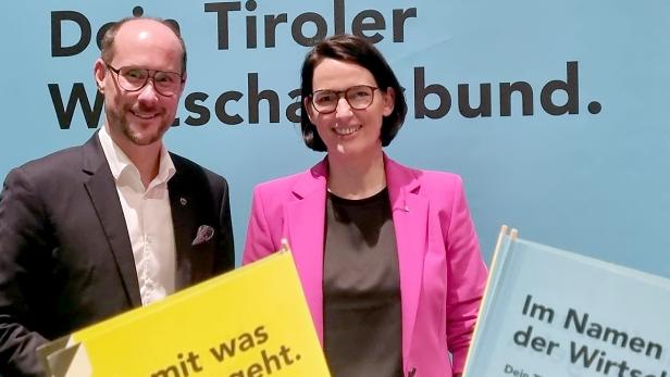 Zwei Personen stehen vor einer blauen Wand mit Schriftzug, beide lächeln und tragen Anzug bzw. pinken Blazer.