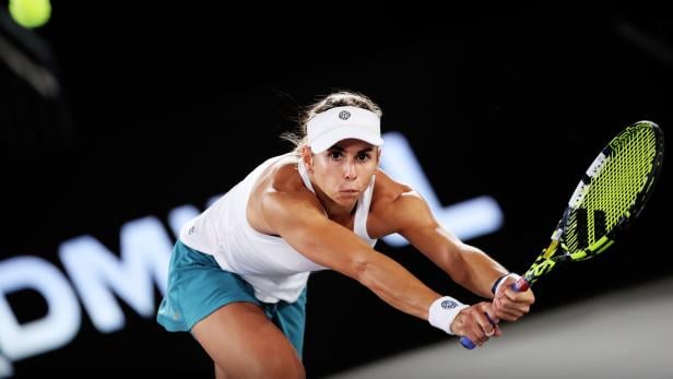 Eine Tennisspielerin in weißem Top und blauem Rock schlägt konzentriert mit beiden Händen einen Ball zurück.