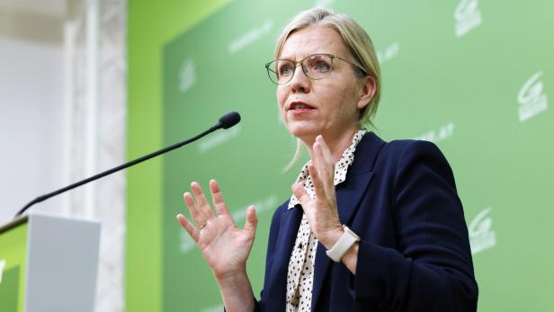 Gewessler gestikuliert vor einem grünen Hintergrund mit Mikrofon.