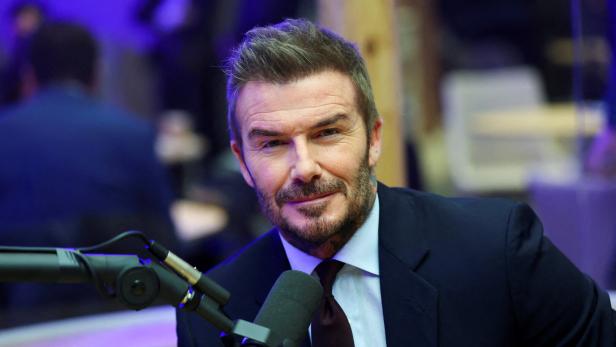 David Beckham lächelt in einem dunklen Anzug vor einem Mikrofon in die Kamera.