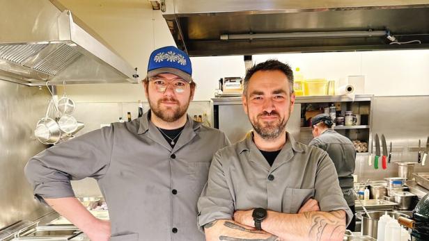 Zwei Köche in grauen Jacken stehen nebeneinander in einer professionellen Restaurantküche.