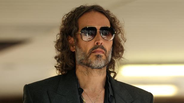 Russell Brand mit lockigem Haar, Bart und Sonnenbrille trägt einen dunklen Anzug und eine Kette mit Anhänger.