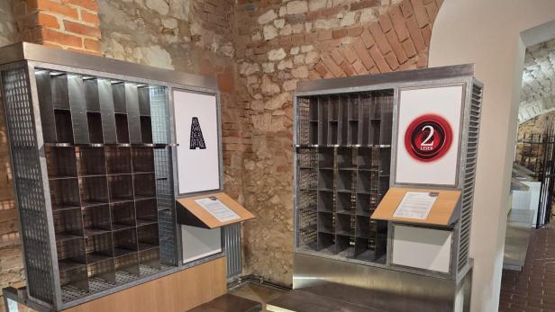Zwei Metallregale mit nummerierten Fächern und Infotafeln stehen vor einer Backsteinwand in einem historischen Raum.