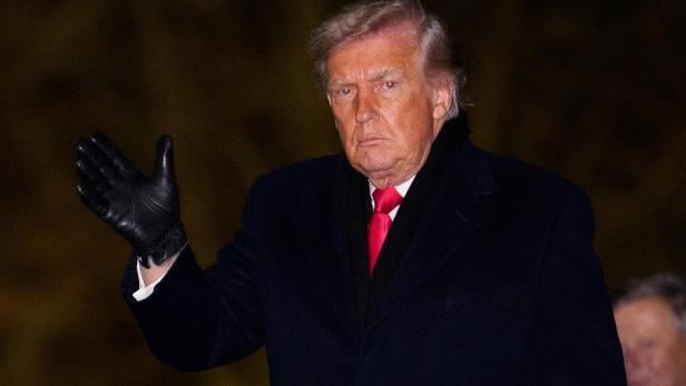 Donald Trump in dunklem Mantel und roter Krawatte grüßt mit einem schwarzen Handschuh.