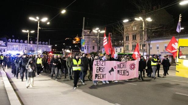Demonstrierende mit Transparenten und roten Fahnen ziehen nachts durch eine Grazer Straße.