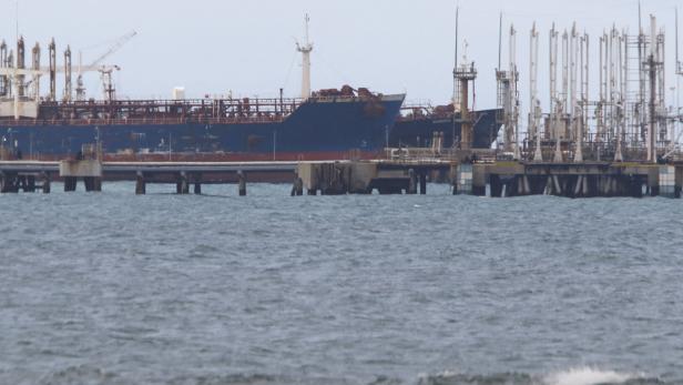 Ein Frachtschiff liegt an einem Industriehafen mit vielen Rohrleitungen und Anlagen am Wasser.