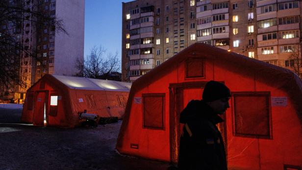Zwei beleuchtete Notfallzelte stehen abends vor einem Wohnblock, eine Person geht im Vordergrund vorbei.