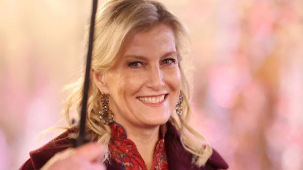 Eine lächelnde Herzogin Sophie mit blonden Haaren trägt einen burgunderroten Mantel und hält eine Kerze und eine Broschüre.