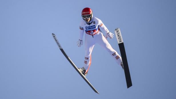 Eine Skispringerin in weißem Anzug und rotem Helm schwebt vor blauem Himmel mit ihren Skiern.