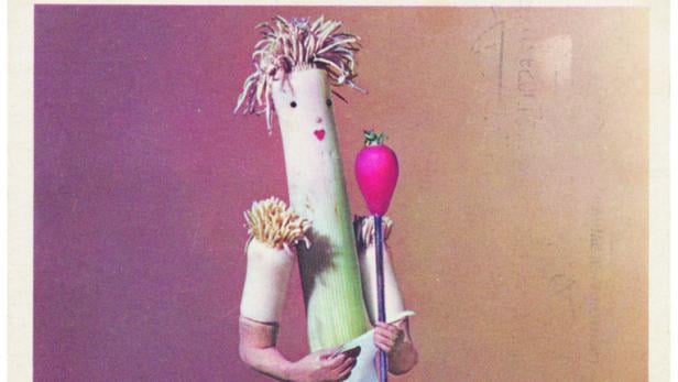 Eine Figur aus Lauch und anderem Gemüse steht auf kariertem Boden und hält ein Mikrofon mit einer roten Kugel.