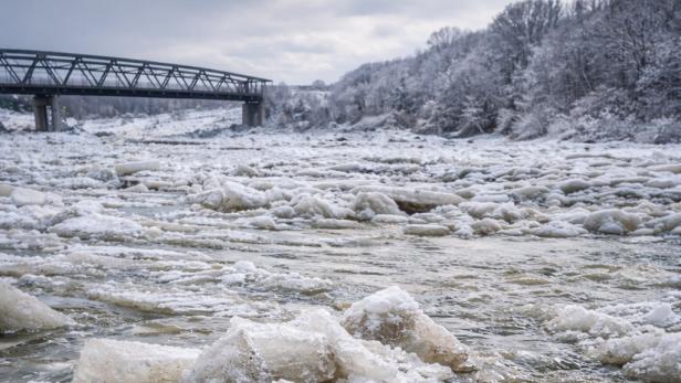 Eine Aufnahmen der von kleinen Eisschollen bedeckten Oberfläche eines Flusses. Im Hintergrund ist eine Brücke erkennbar.