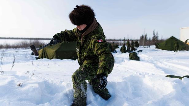 Eine Person in Wintertarnkleidung schlägt mit einem Hammer in den Schnee, im Hintergrund weitere Personen und Zelte.